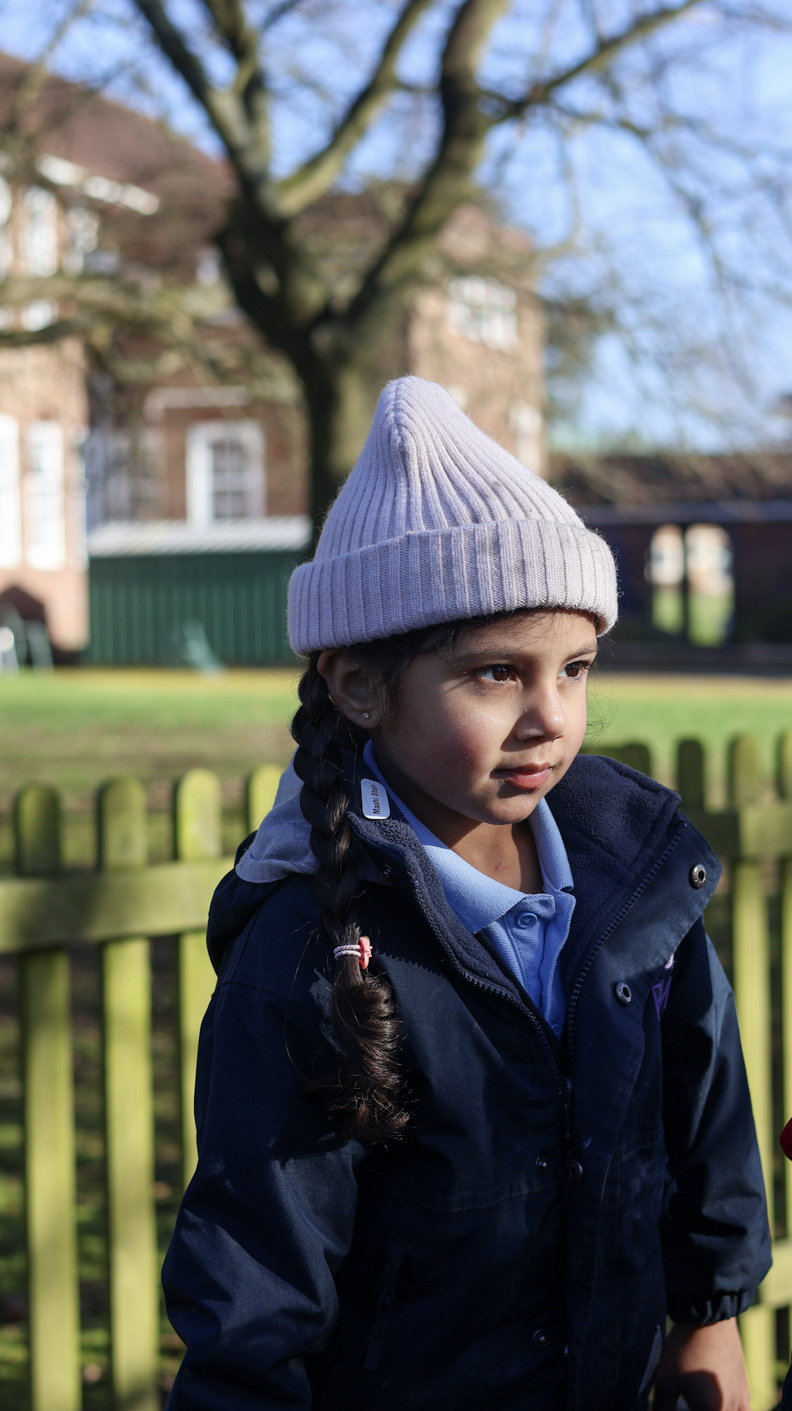 Young girl wearing a light gray knit beanie and navy jacket outdoors, with a blurred brick building and trees in the background.