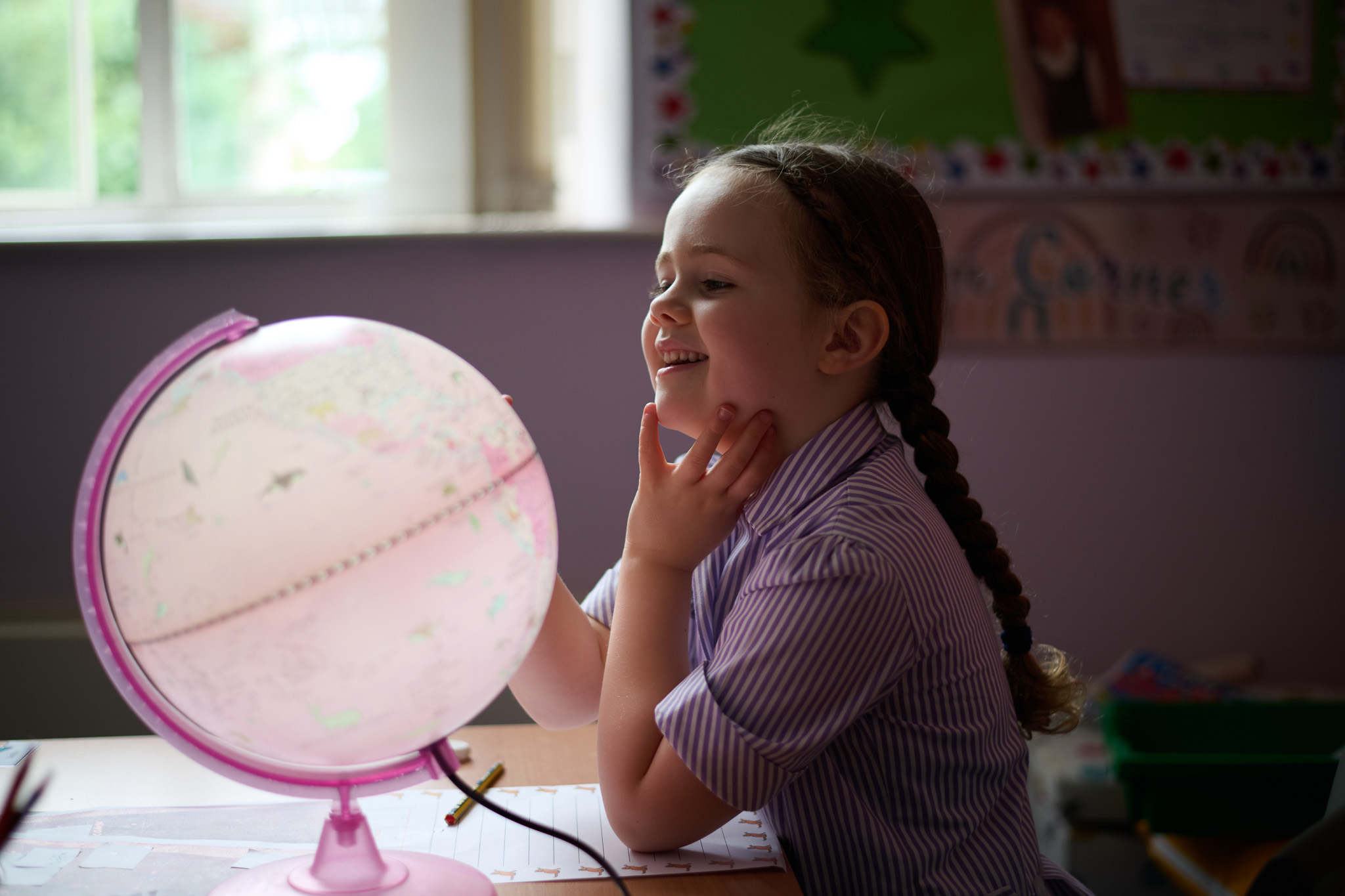 A smiling girl in a striped shirt studies a pink globe at a desk in a classroom.