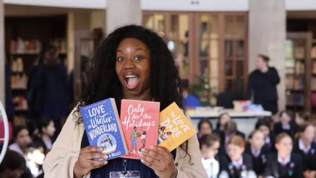Excited woman holding three colorful children's books at a library event with a blurred audience in the background.