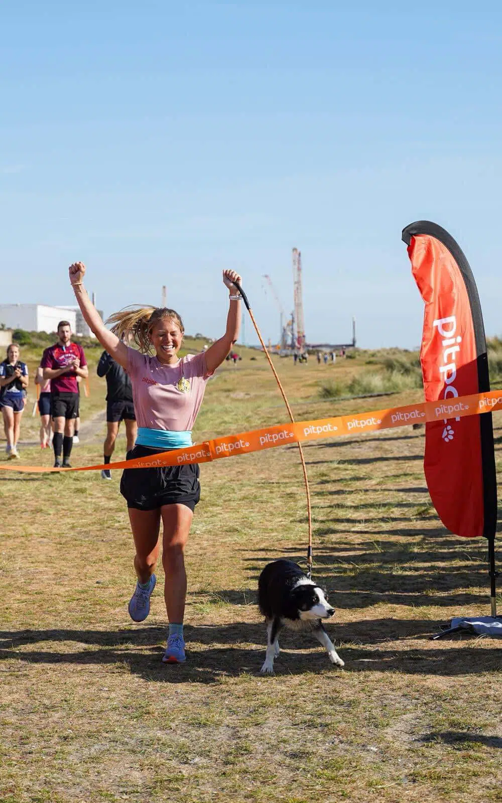 Female runner with arms raised crosses a finish line as a black-and-white dog trots beside her, orange 'pitpat' tape marking the line under a blue sky.