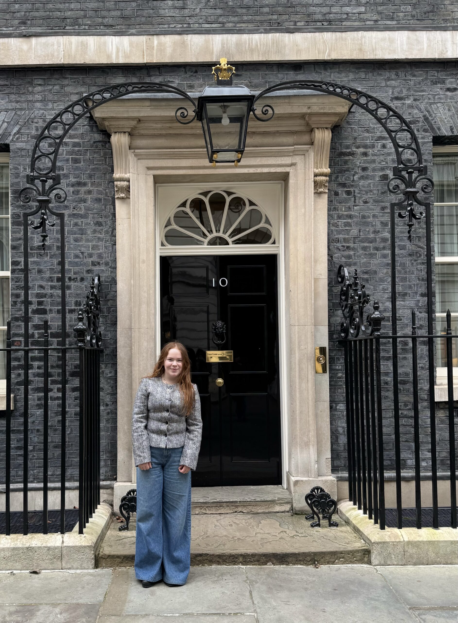 Girl standing on steps in front of a black front door labeled with the number 10, framed by a beige stone doorway and black wrought-iron gates against a gray brick facade.