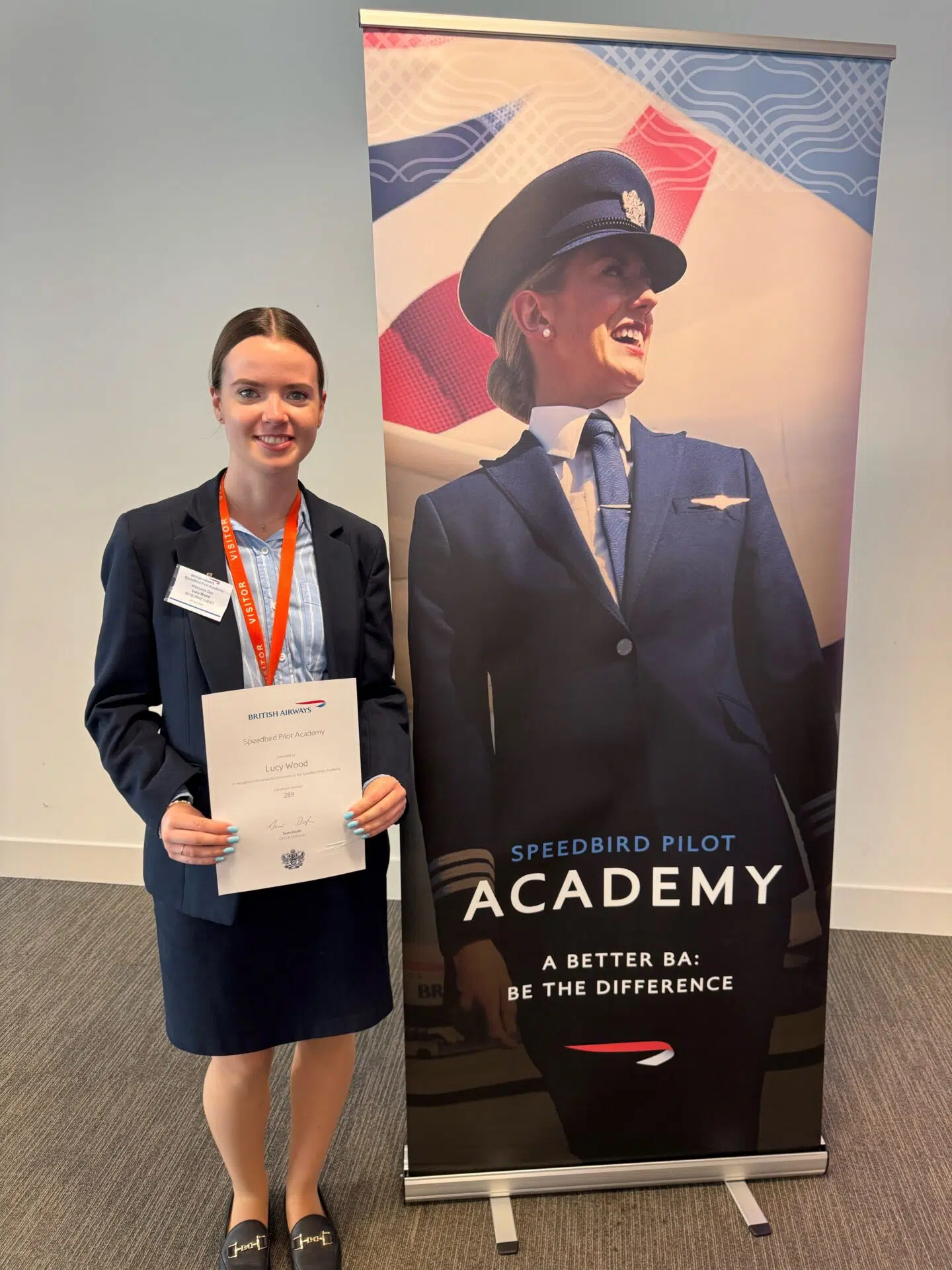 Young woman in a navy blazer holds a certificate beside a Speedbird Pilot Academy banner featuring a smiling pilot in uniform.
