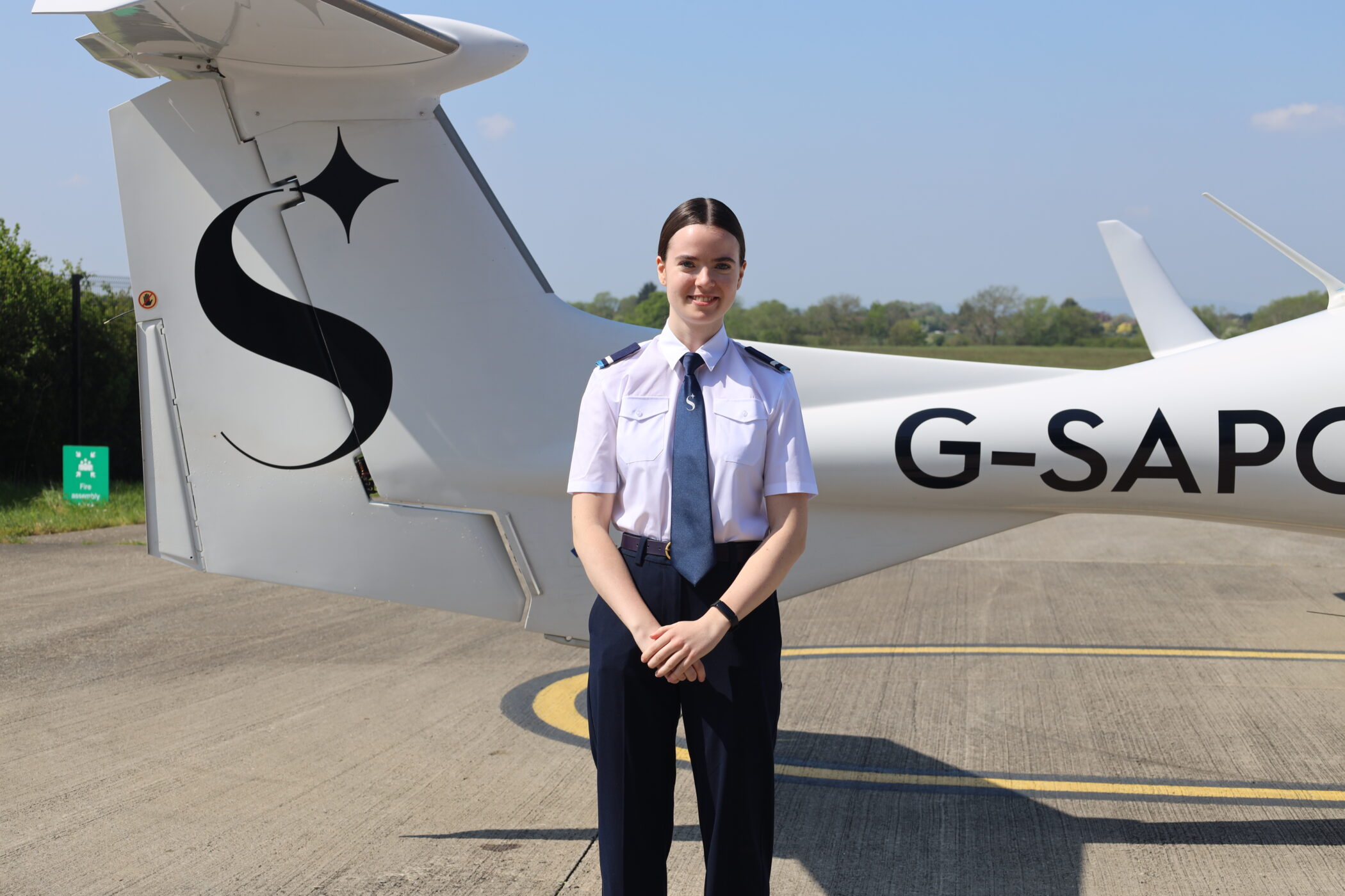Female pilot in uniform stands beside a white airplane on a tarmac.