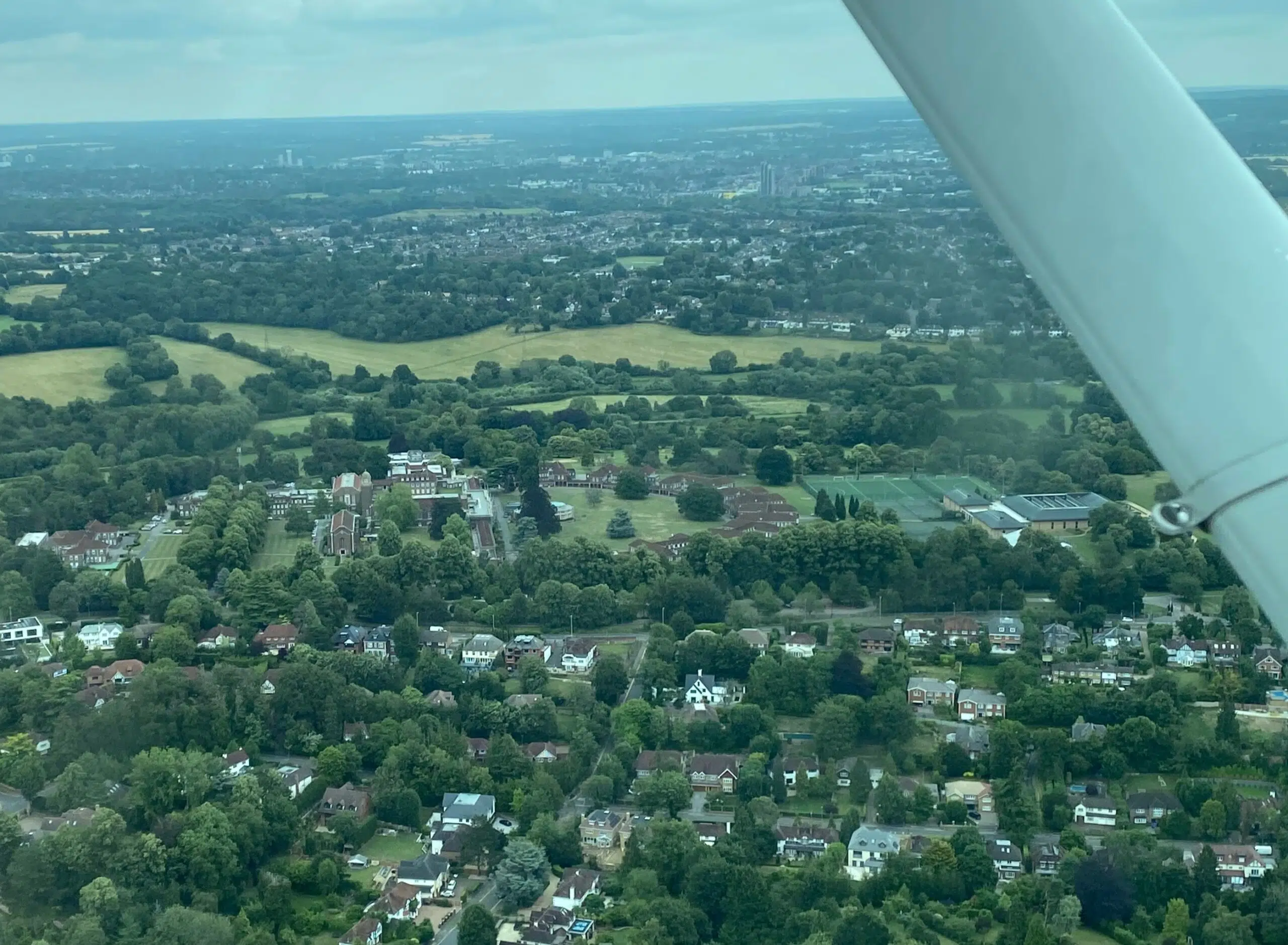 Aerial view over a suburban town with houses, trees, and open fields; an airplane wing is visible along the right edge.