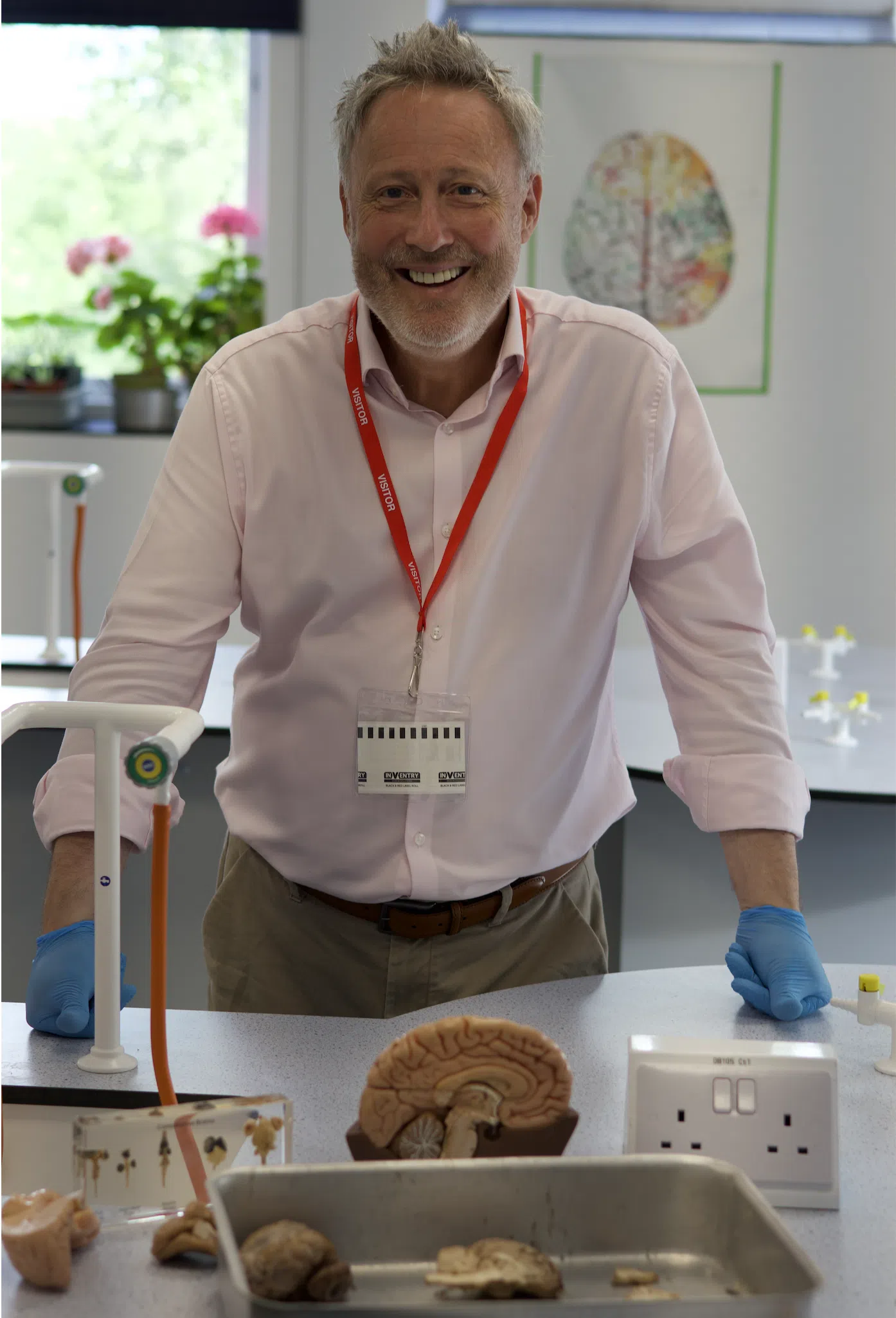 Smiling man in a pink shirt with a red visitor badge stands at a lab table with brain models and dissection trays in a classroom lab setting.
