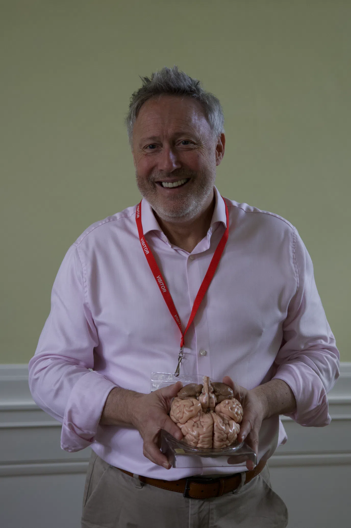 Smiling man in a pink shirt with a red visitor badge holding a glass dish containing a brain model.