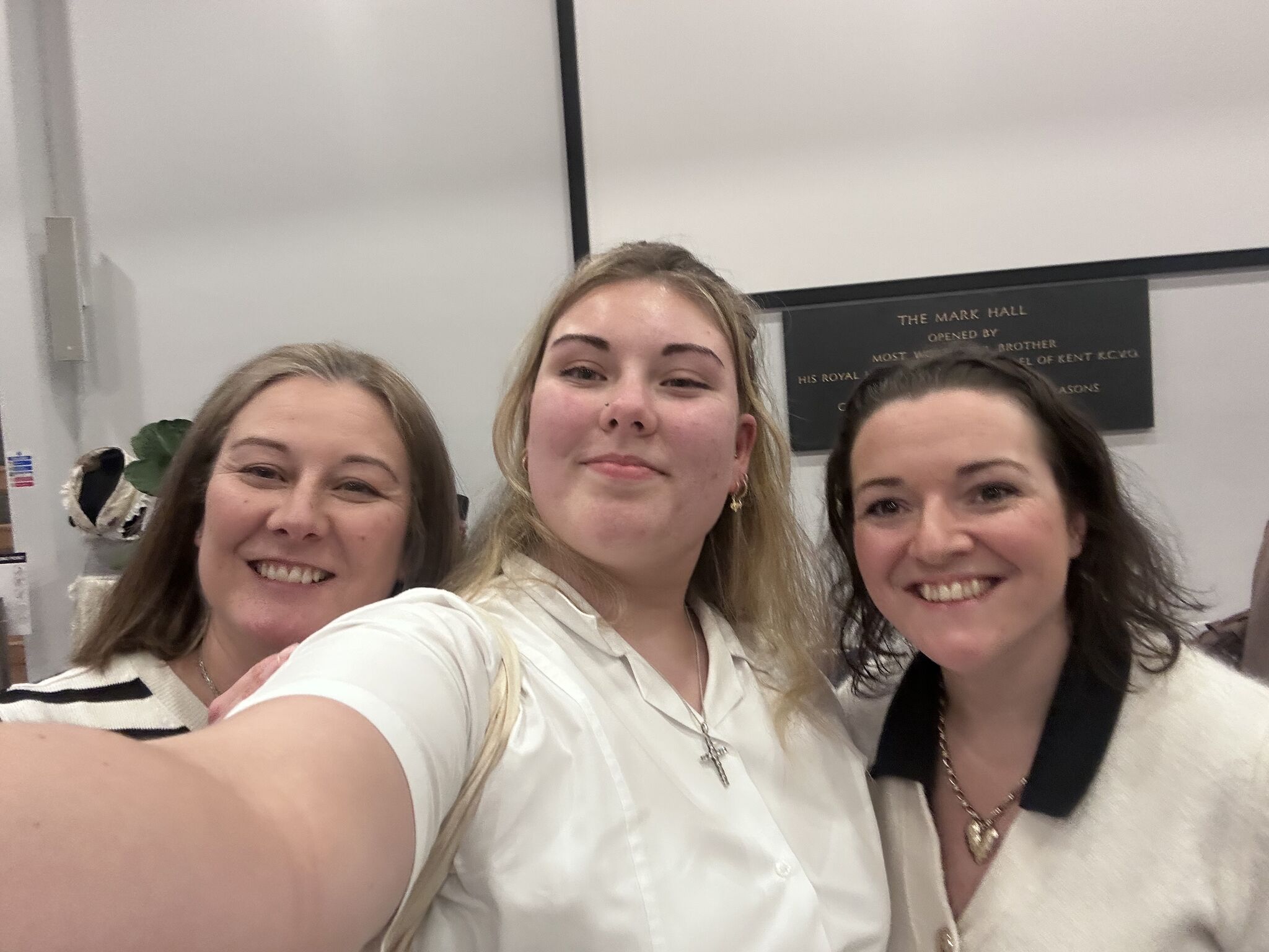 Three smiling women taking a selfie indoors, wearing white tops.