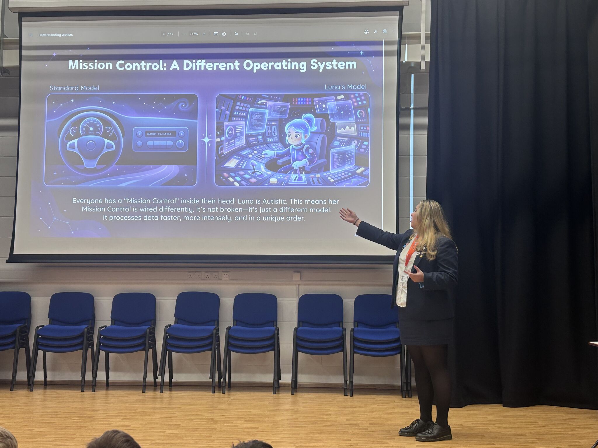 Presentation slide titled 'Mission Control: A Different Operating System' comparing Standard Model and Luna's Model; presenter gestures at right, blue auditorium chairs in front.