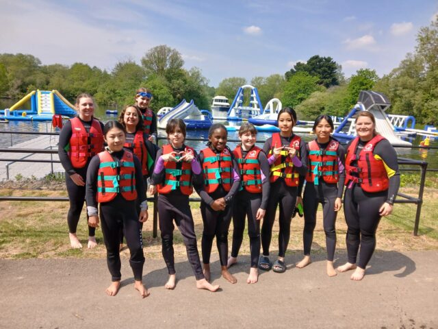 Group of people in red life jackets standing on a path by a lake with inflatable water obstacles in the background, smiling at the camera.