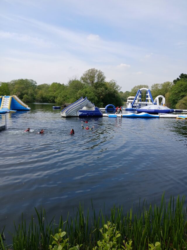 People swimming and playing on large blue and white inflatable water obstacles on a calm lake with trees and blue sky in the background.