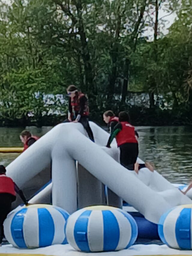 People wearing red life jackets on a boat with blue-and-white striped flotation buoys on a river, trees in the background.