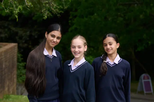 Three girls in matching school uniforms smiling at the camera outdoors.