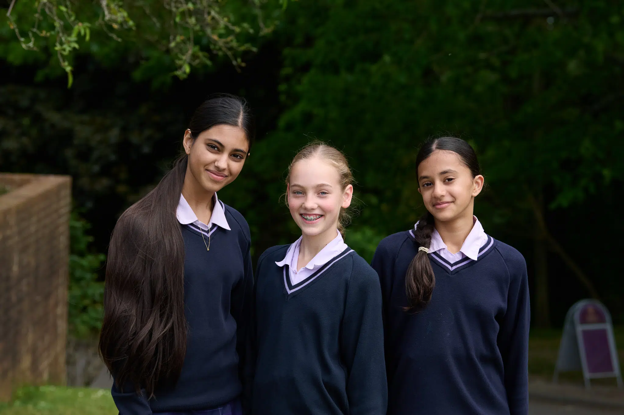 Three girls in matching school uniforms smiling at the camera outdoors.