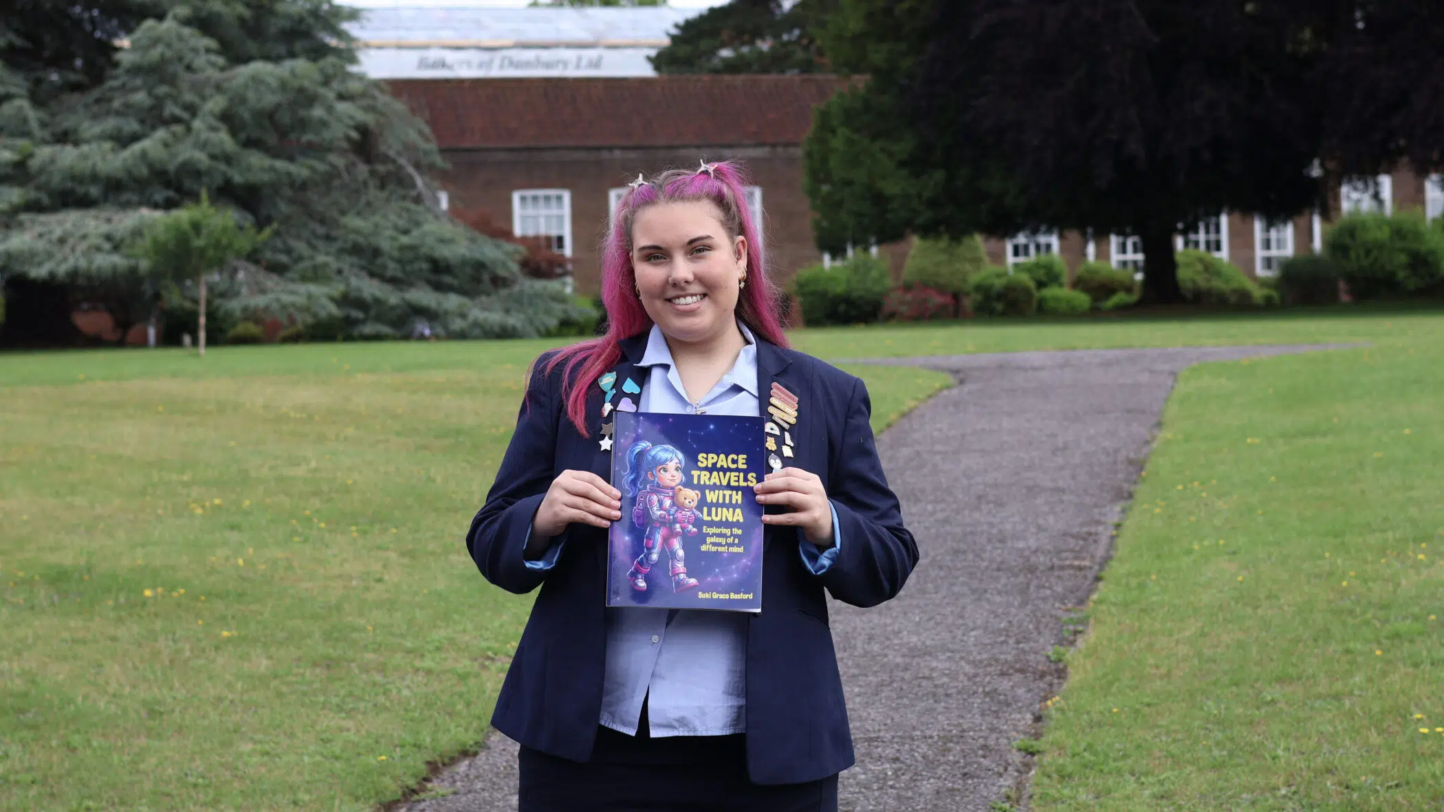 Young woman with pink hair holding a Space Travels with Luna book on a grassy campus path, smiling at the camera.