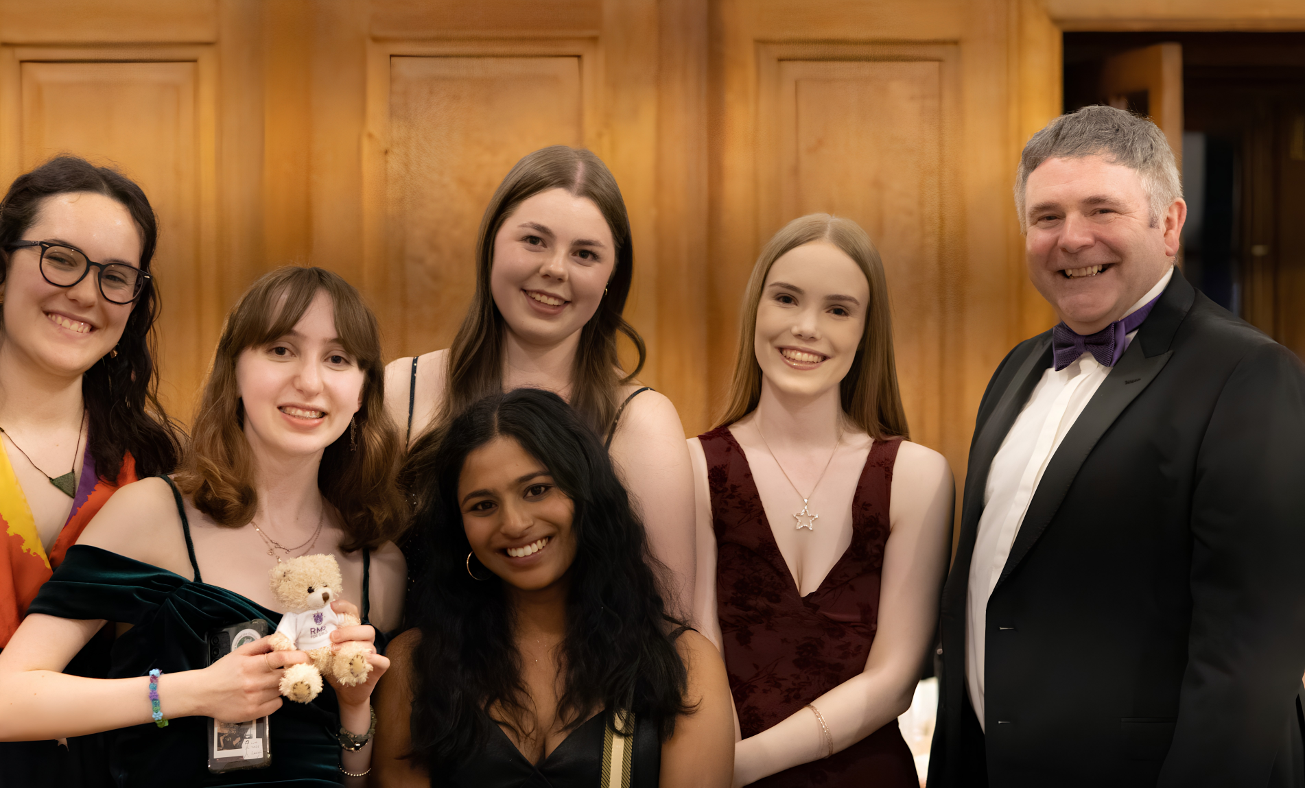 Smiling group of six in formal attire posing for a photo indoors, with a wooden paneled wall in the background. A man in a tuxedo stands on the right.
