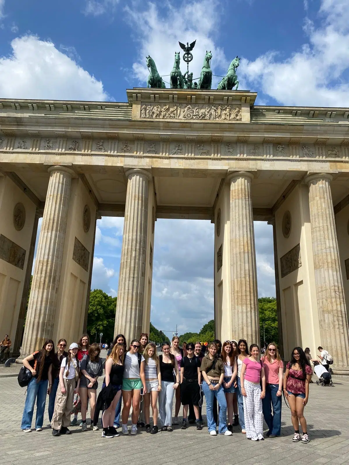 Group of young tourists posing in front of a grand neoclassical arch with statues on top and tall columns, blue sky above.
