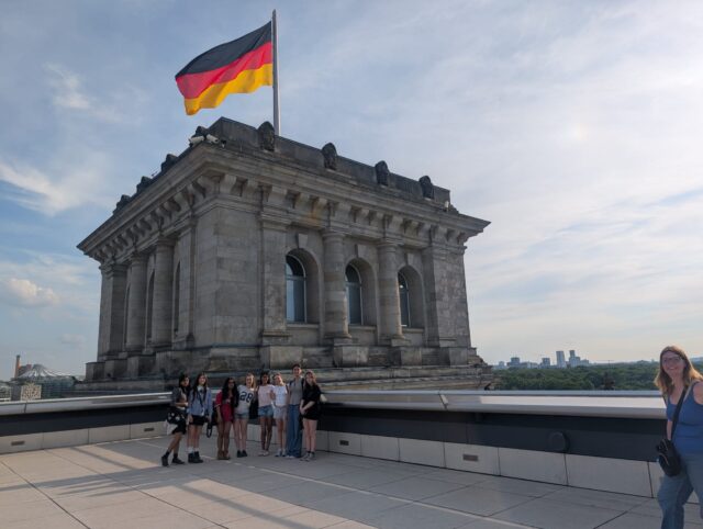 Group of people posing on a viewing platform beside a neoclassical stone building with a tall German flag overhead.