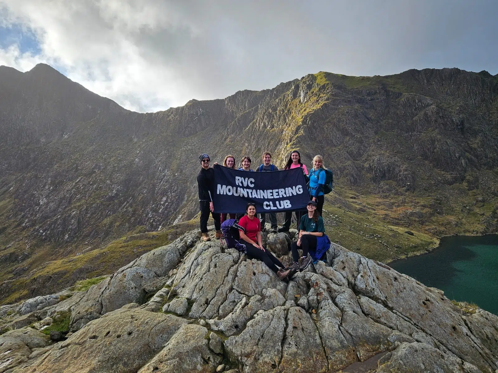 Group of hikers on a jagged mountain summit holding a banner that reads 'RVC MOUNTAINEERING CLUB' with rocky slopes and a lake in the background.