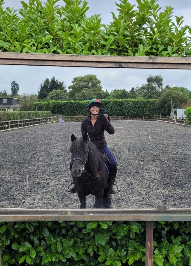 A smiling rider wearing a helmet on a dark horse, taking a selfie in an outdoor riding arena with green hedges in the background.