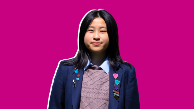 Portrait of a young girl in a navy blazer with colorful badges, smiling at the camera against a magenta background.
