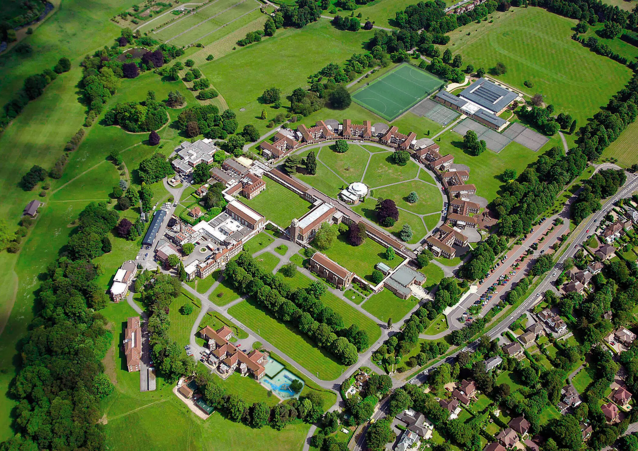 Aerial view of a university campus with green fields, a soccer field, tennis courts, and cluster of red-brick buildings connected by curved paths.
