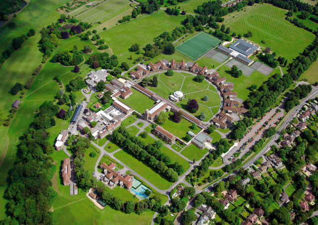 Aerial view of a university campus with green fields, a soccer field, tennis courts, and cluster of red-brick buildings connected by curved paths.
